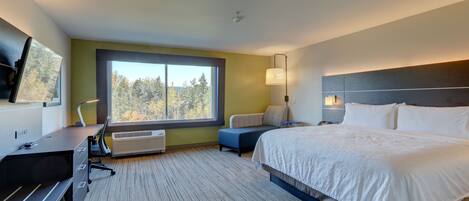A modern hotel room with a large bed, a desk, and a view of trees through a window.