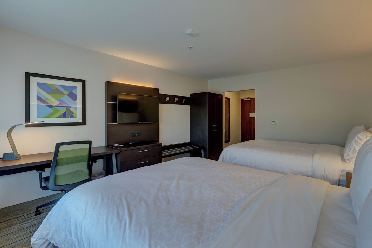 A hotel room with two beds, a desk with a chair, a dresser, a mirror, and a framed picture on the wall.