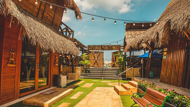 A courtyard with thatched-roof buildings, a wooden bench, and a string of lights. - Imagine Beach (Puerto Colombia)