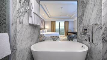 A modern bathroom with a large bathtub, marble walls, and a view of a bedroom with a bed and curtains.