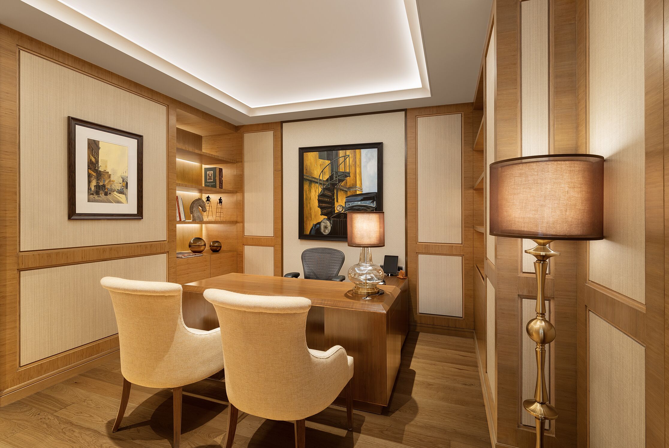 A modern hotel room with a wooden desk, two chairs, a lamp, and framed artwork on the walls.