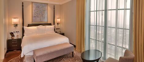 A hotel room with a large bed, a bench, a round table, and a chair. There is a large window with curtains, a painting on the wall, and a nightstand with a lamp.