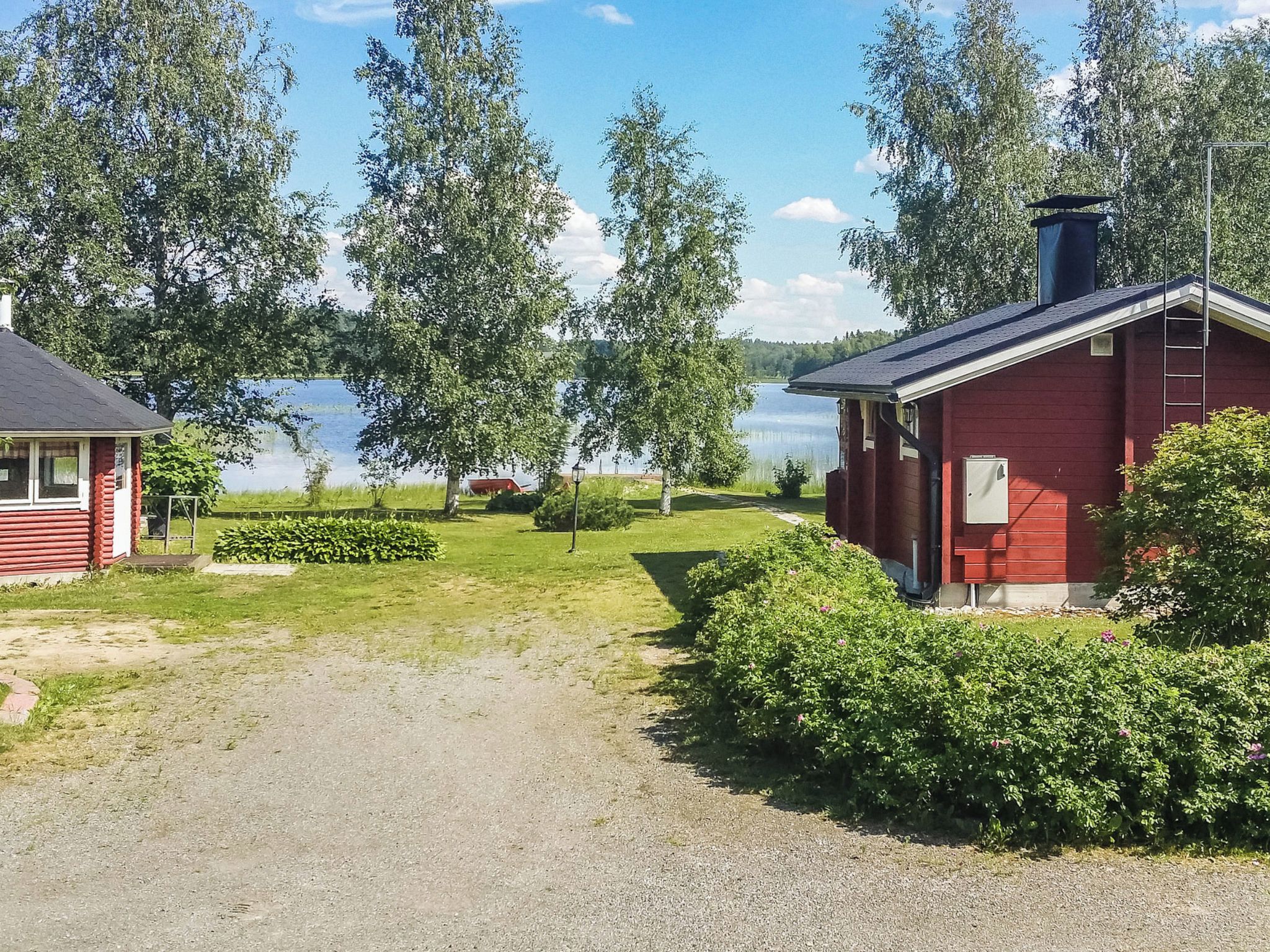 Punainen mökki, jossa on savupiippu, ja sen ympärillä on vehreää kasvillisuutta sekä kivituhkapolku, joka johtaa järvelle.
