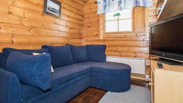 L’intérieur d’un chalet en bois, avec un canapé d’angle, un téléviseur et une fenêtre avec des rideaux.