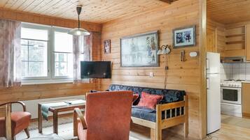Interior acogedor de una cabaña con suelo de madera, un sofá, una silla, una mesa de comedor, un televisor y una cocina con estufa y refrigerador.