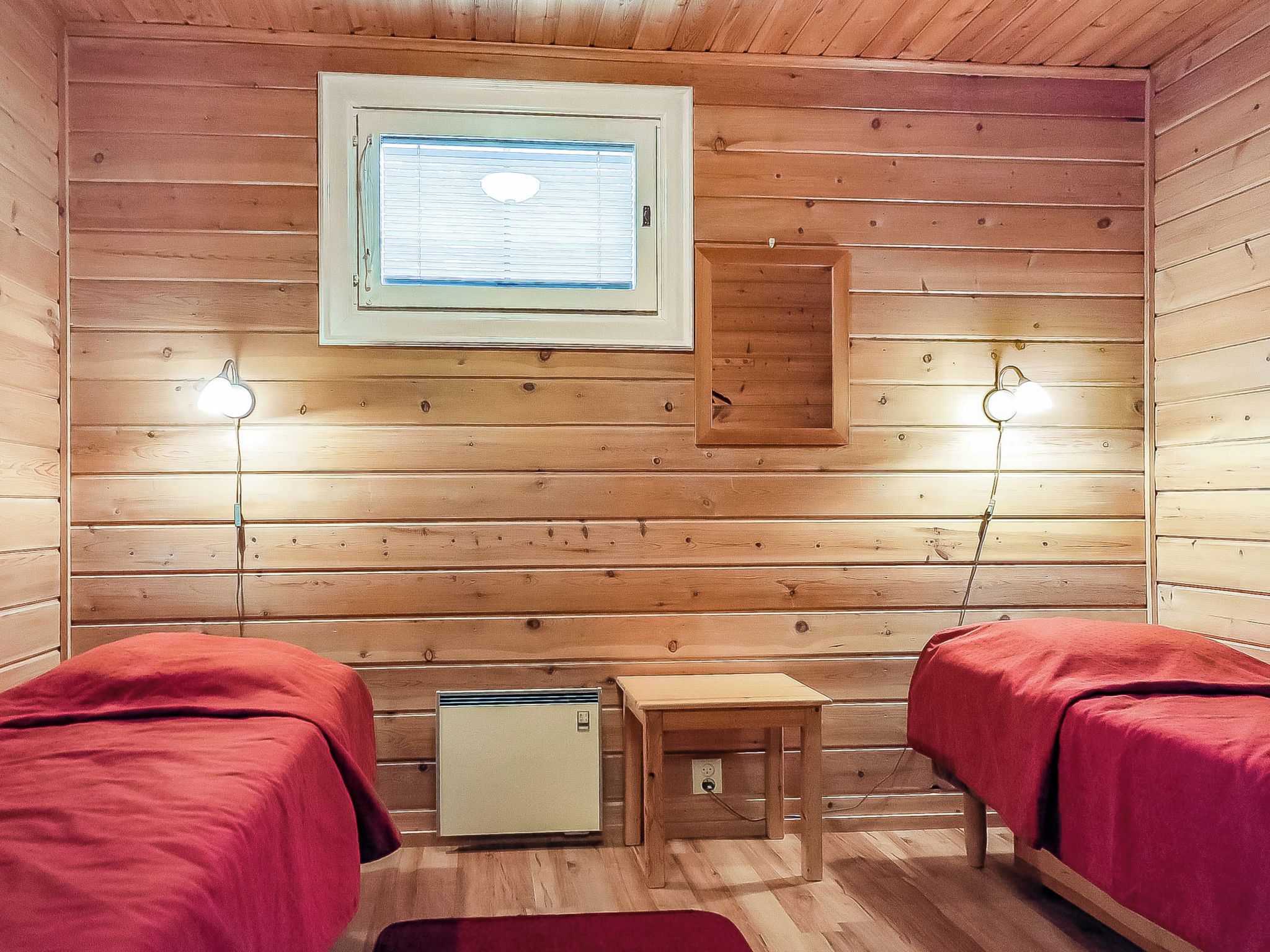 A small, cozy room with wooden walls, two beds with red bedding, a small table, a heater, and two wall-mounted lamps.