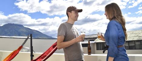 A man and a woman standing on a rooftop terrace with a mountain in the background, holding drinks.