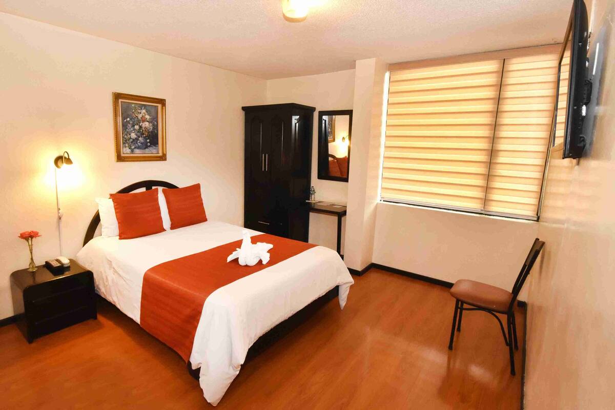 Une chambre d’hôtel avec un lit, un bureau, une chaise, une table de chevet, une lampe, un tableau et une fenêtre avec des stores.