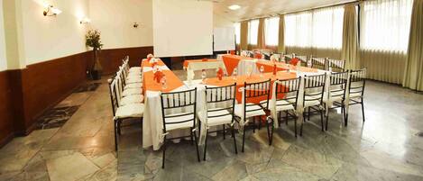 Un salón de conferencias con una larga mesa y sillas, una pantalla de proyector y una planta en maceta.