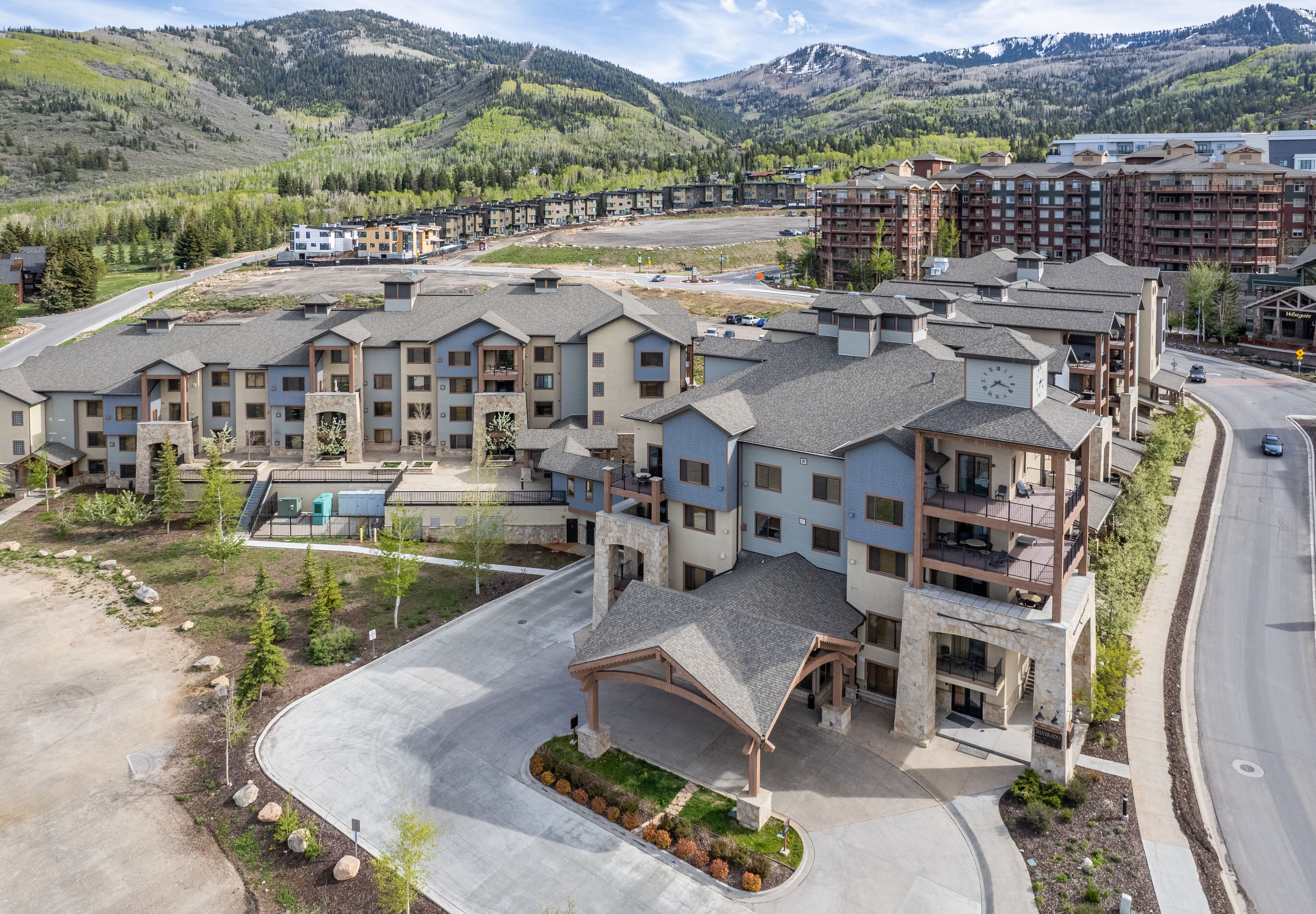 A modern hotel complex with multiple buildings, a covered parking area, and a view of surrounding mountains.