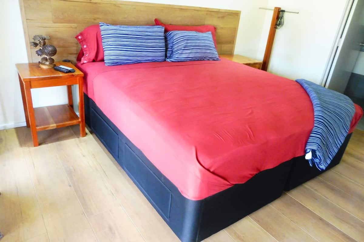 A bed with a red bedspread and two striped pillows, a wooden nightstand with a remote control, and a small decorative item.