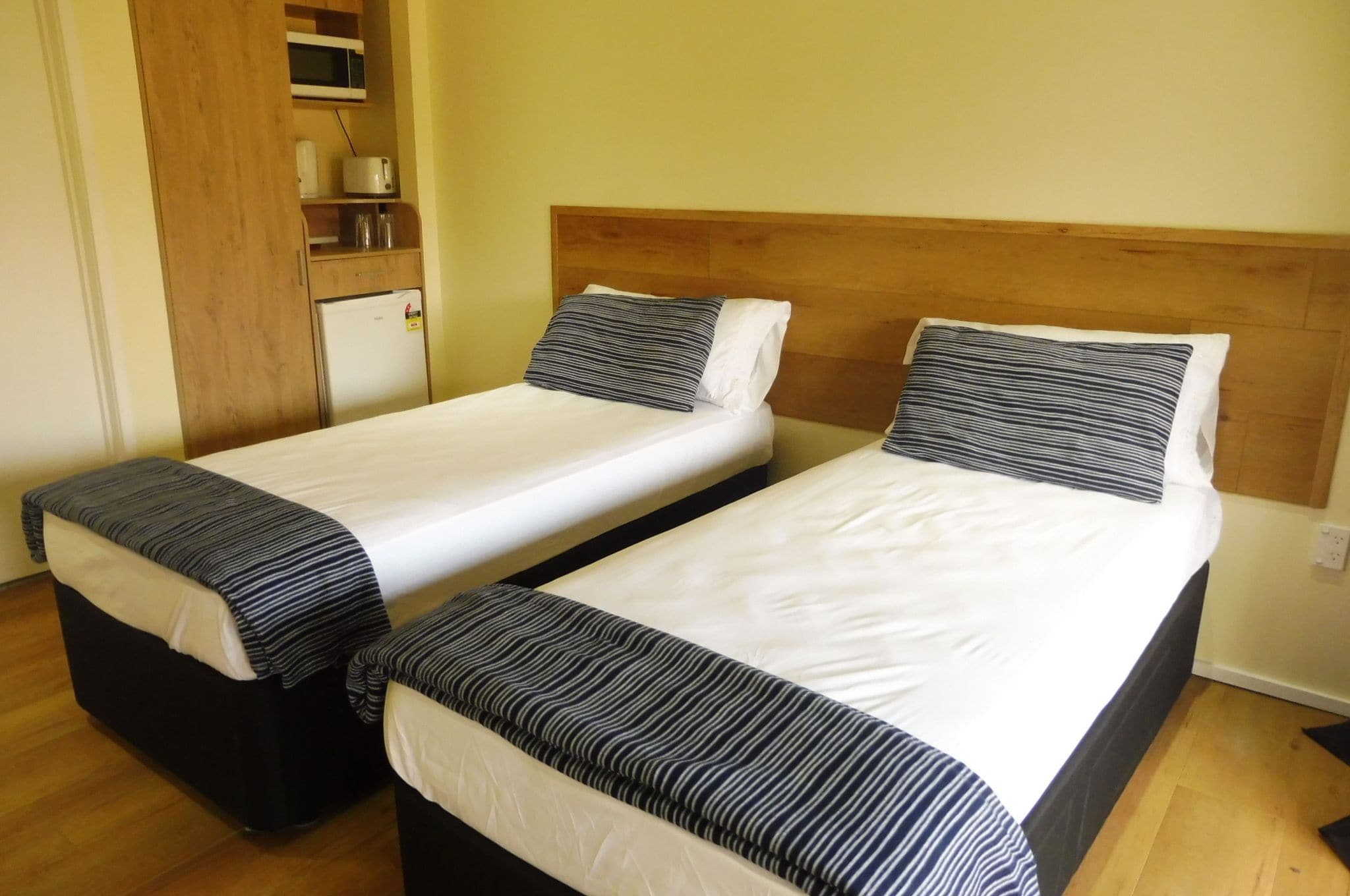 Dos camas individuales con ropa de cama a rayas, un cabecero de madera, una pequeña kitchenette con microondas y un armario de madera.
