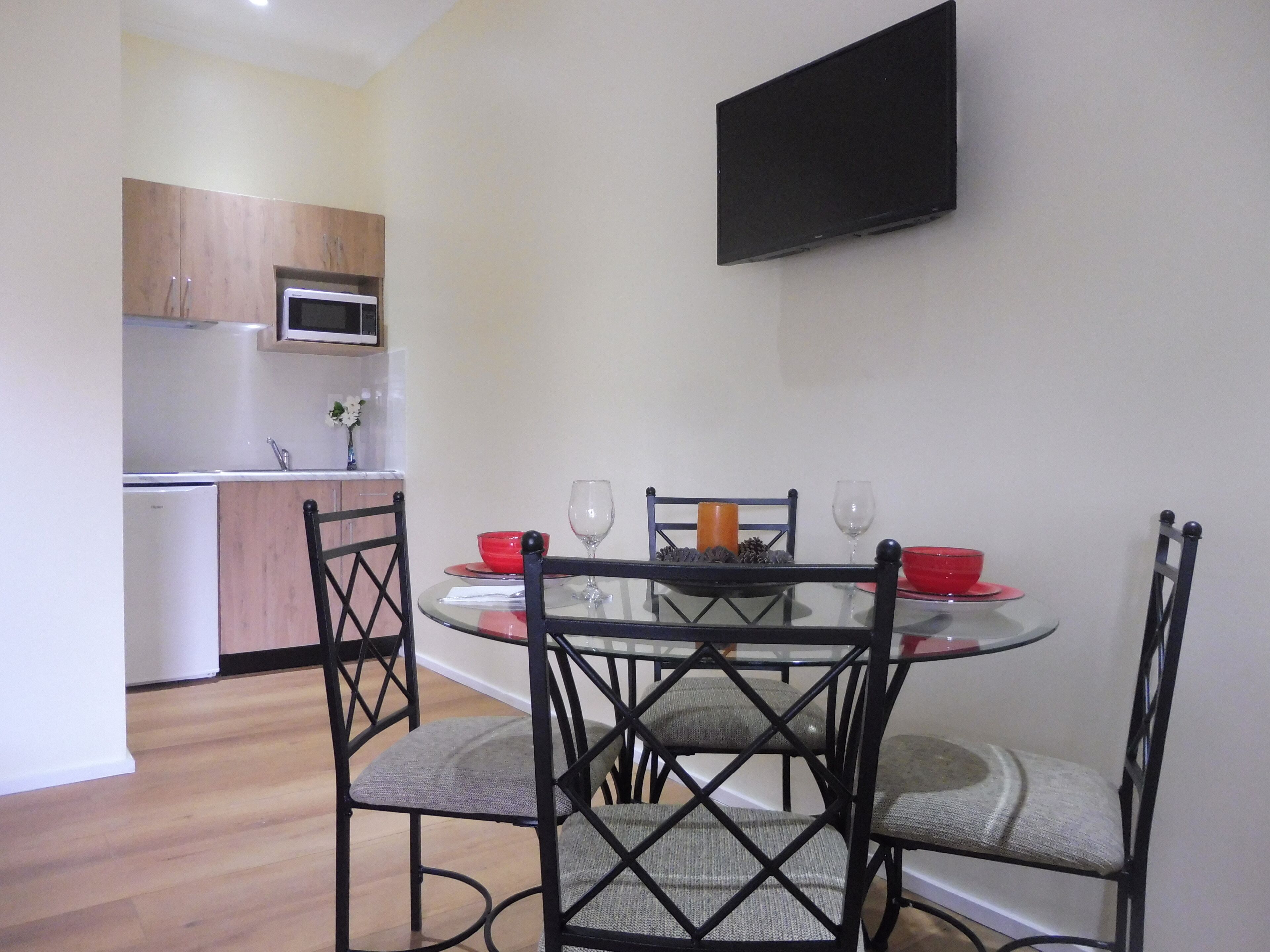 Una cocina compacta con electrodomésticos modernos, un comedor con mesa de vidrio y sillas de metal, y un televisor de pantalla plana montado en la pared.