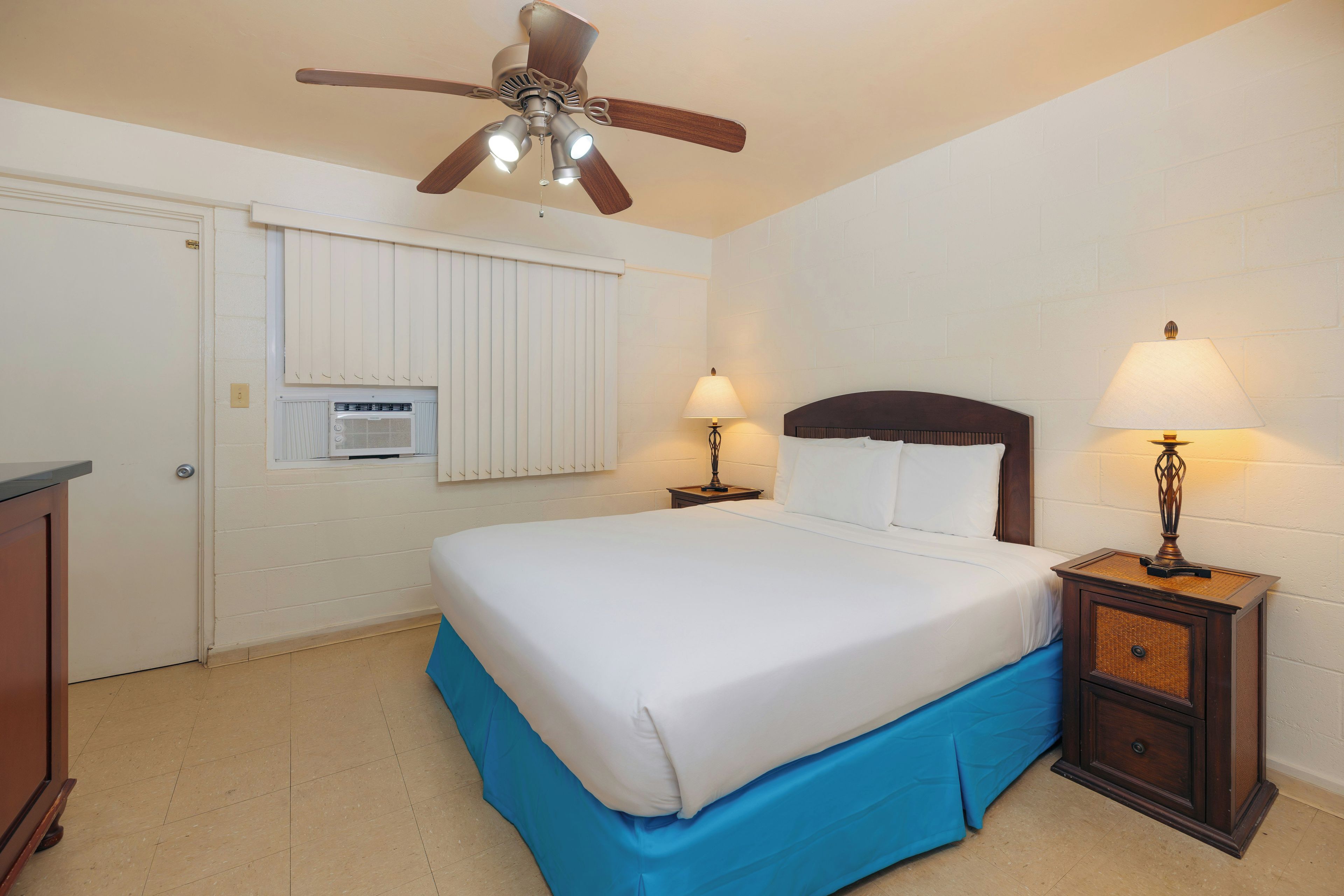 A bedroom with a bed, two bedside lamps, a ceiling fan, a window with blinds, and a radiator.