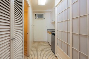 A narrow hallway with a tiled floor, a door with a handle, a white air conditioning unit mounted on the wall, and a tiled wall with a grid pattern. - Waikiki Heritage Hotel (Honolulu)