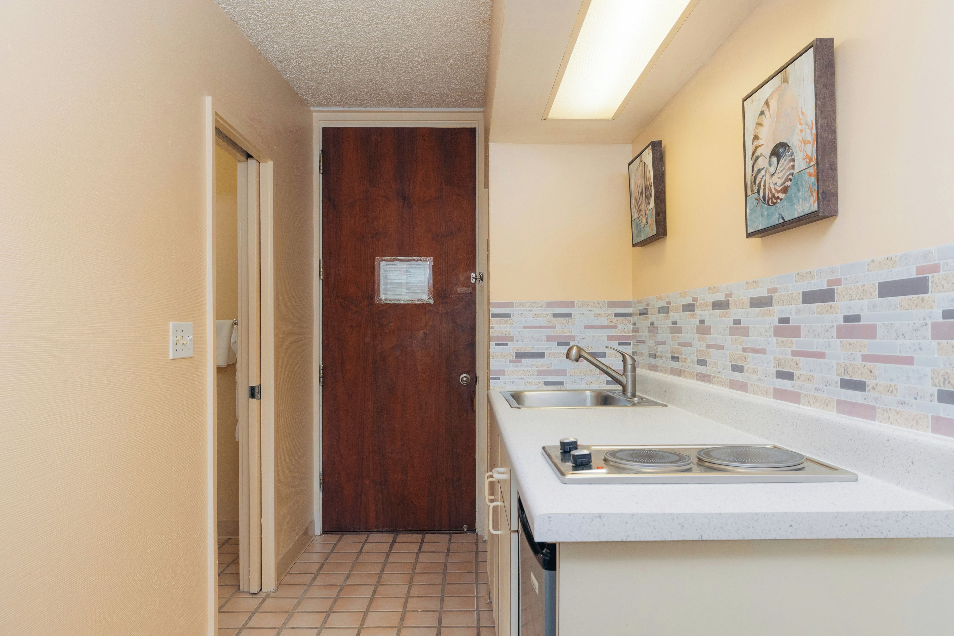 Un pasillo estrecho que lleva a una puerta de madera, una cocina con salpicadero de azulejos y un lavamanos.