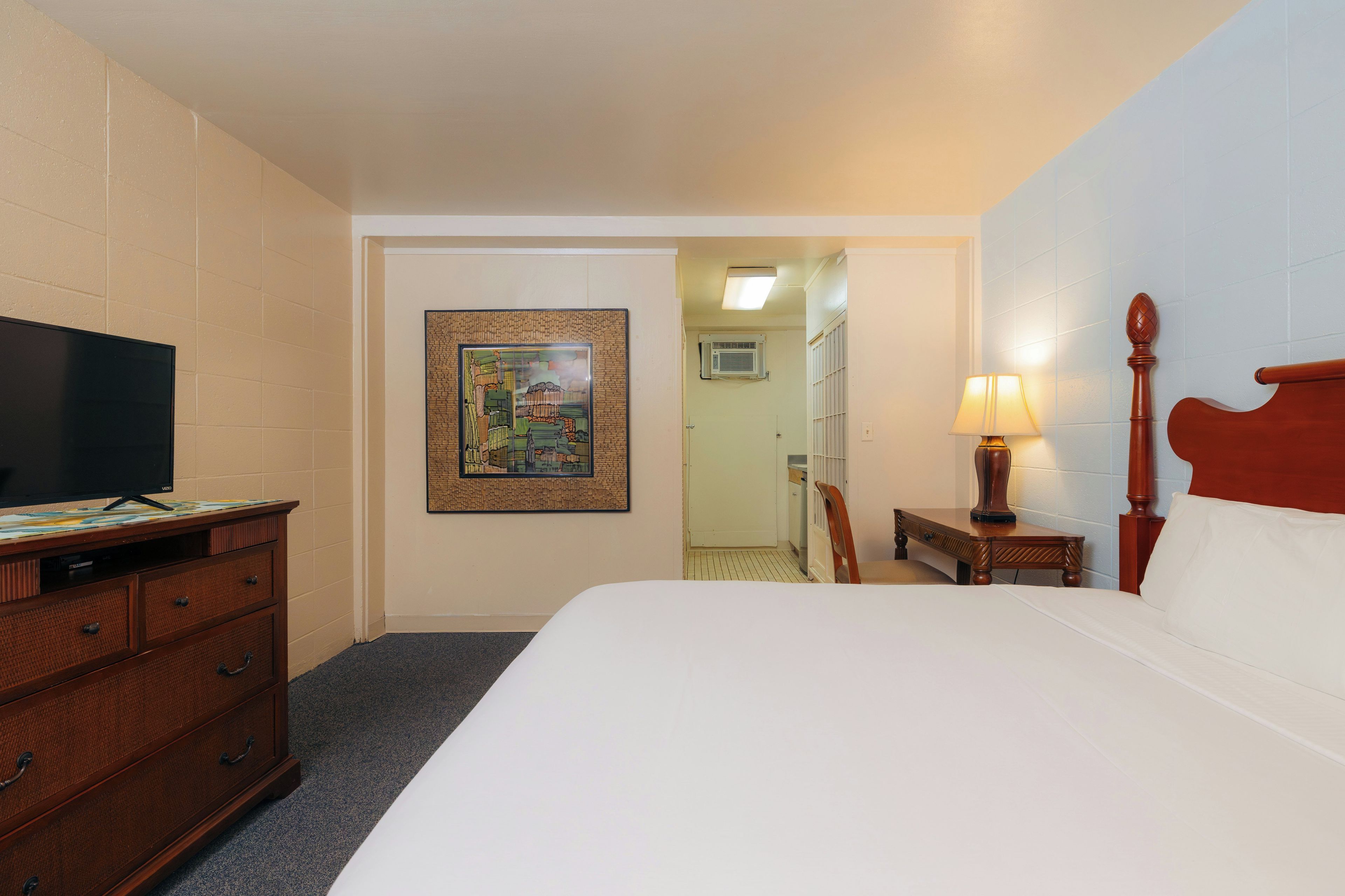 Quarto de hotel com cama, televisão, escrivaninha com gavetas, um quadro na parede, luminária e uma cadeira.