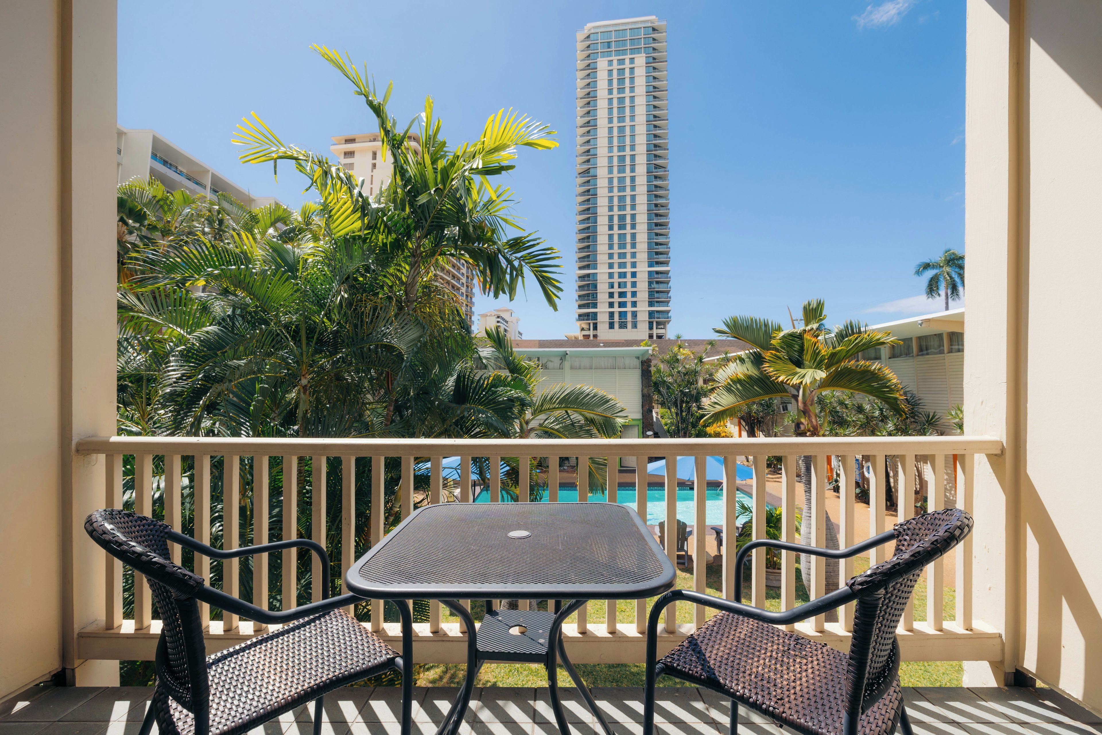 Un balcón con mesa y sillas, con vistas a una piscina y palmeras, con un edificio alto al fondo.