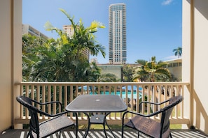 A balcony with a table and chairs, overlooking a pool and palm trees, with a tall building in the background. - Waikiki Heritage Hotel (Honolulu)