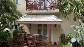 Un edificio de dos pisos con un patio cubierto, un balcón con baranda de madera y un pequeño jardín con diversas plantas.