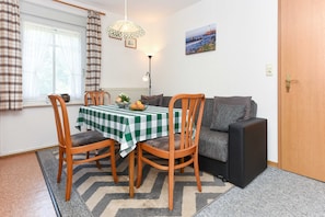 A cozy room with a dining table set for two, a sofa, a black speaker, and a painting on the wall.