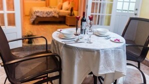 Une table à manger dressée pour deux personnes, avec une nappe blanche, deux chaises, et en arrière-plan, une vue sur une chambre à coucher.
