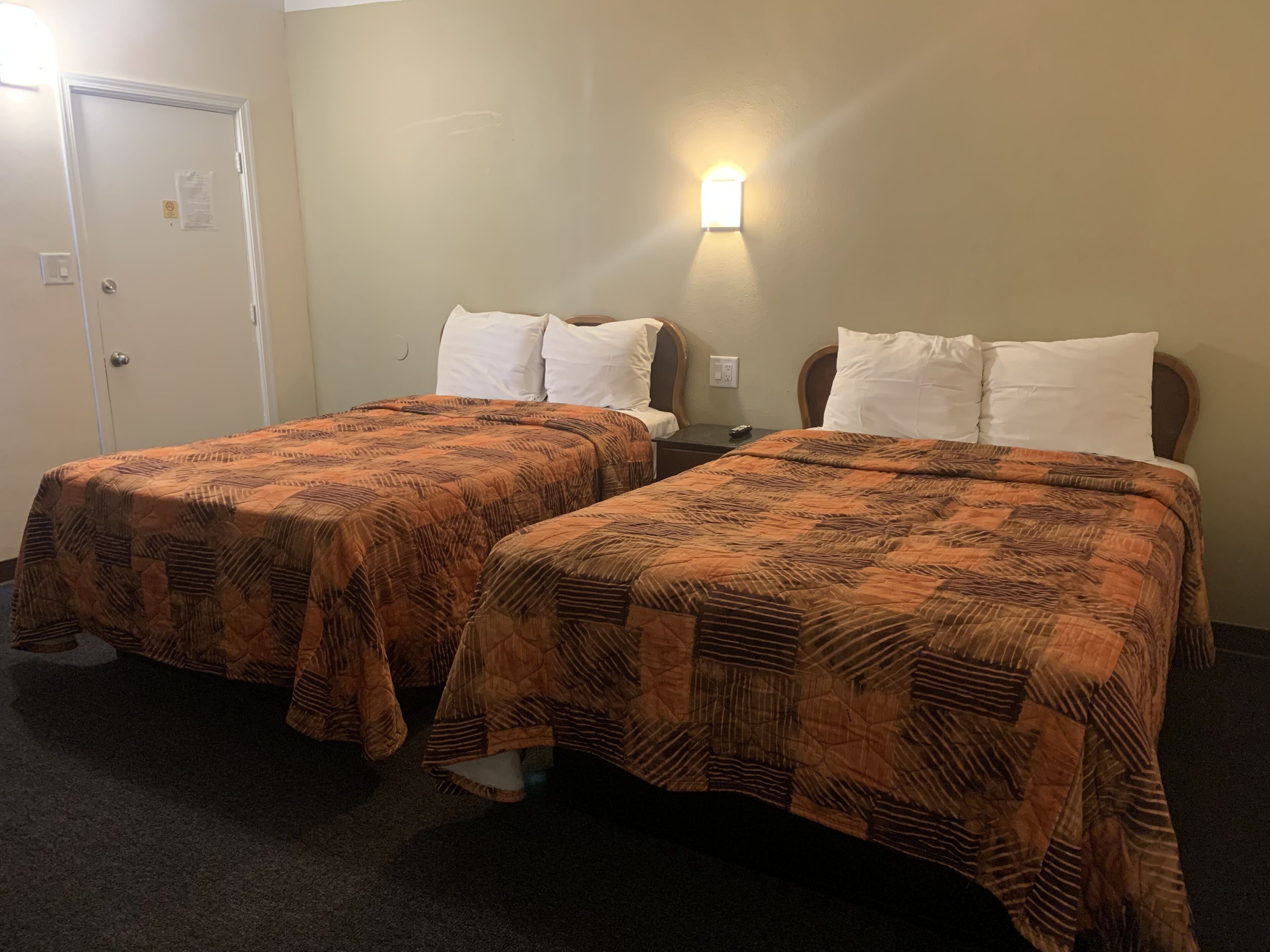 A hotel room with two beds, each with a patterned bedspread and white pillows. There is a nightstand between the beds with a lamp and a remote control. The room has a carpeted floor and a door to the left.