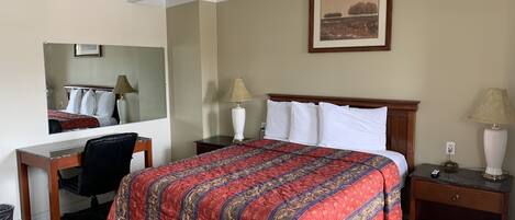 A hotel room with a large bed, a desk, a chair, a nightstand, a lamp, a mirror, and a framed picture on the wall.