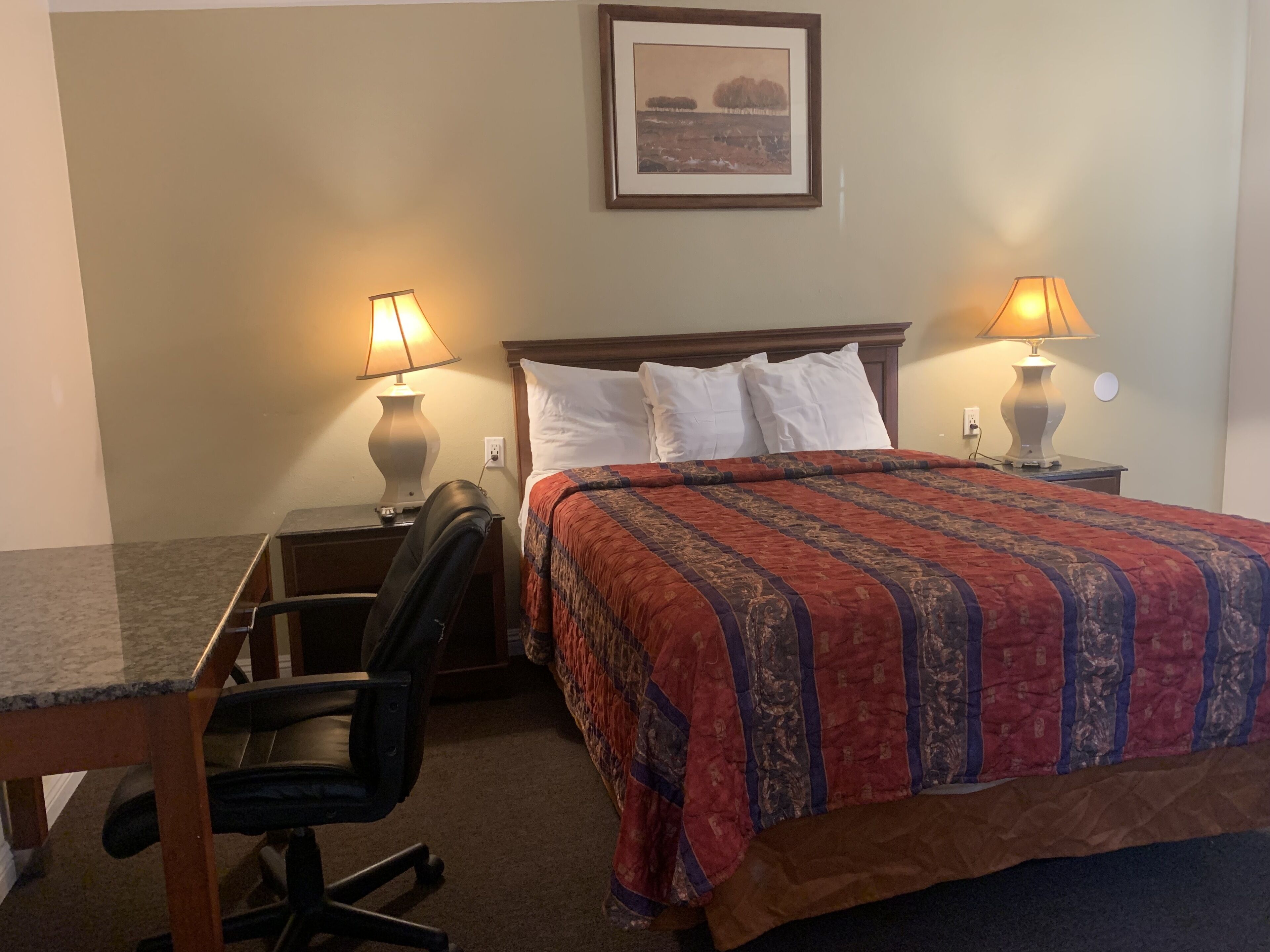 Una habitación de hotel con una cama, dos lámparas de noche, un escritorio con una silla y un cuadro en la pared.