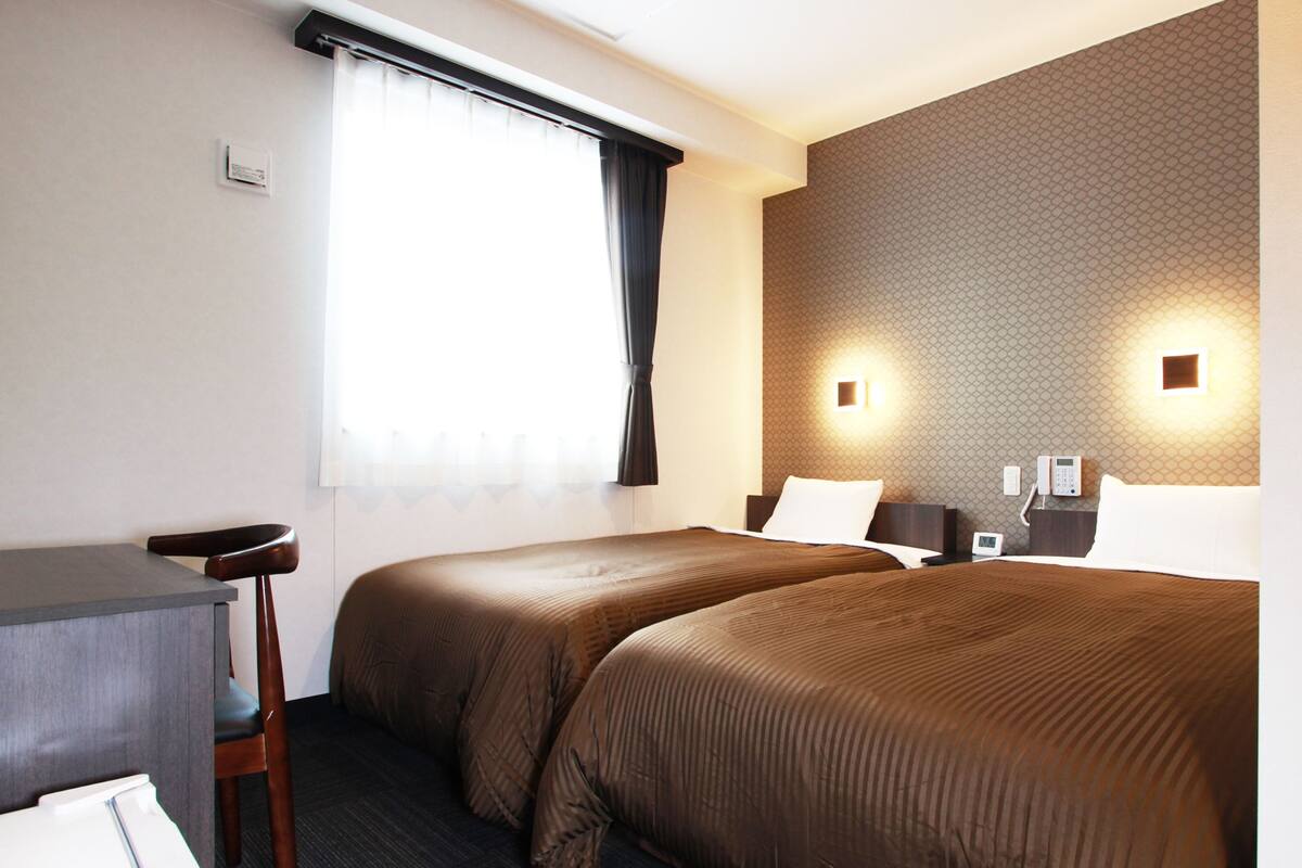 Habitación de hotel con dos camas, un escritorio, una silla, un ventanal con cortinas y un papel tapiz estampado.