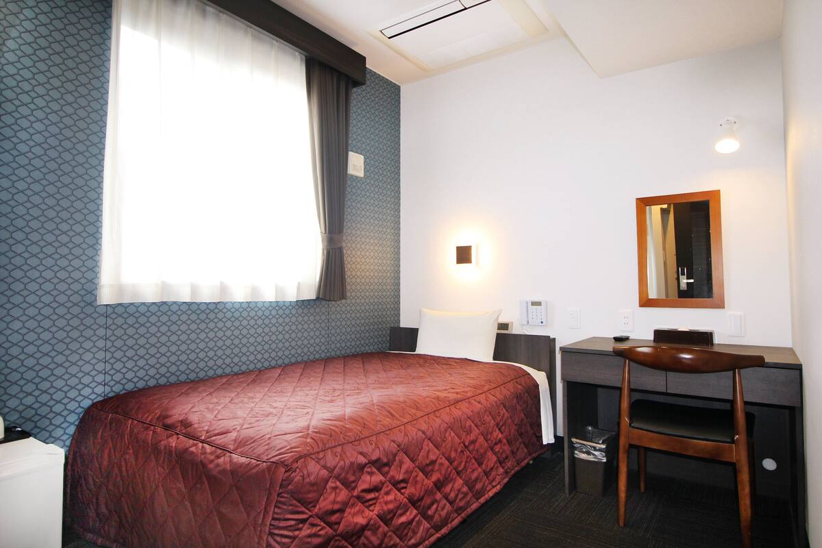 Habitación de hotel con cama, escritorio, silla, espejo y ventana con cortinas.