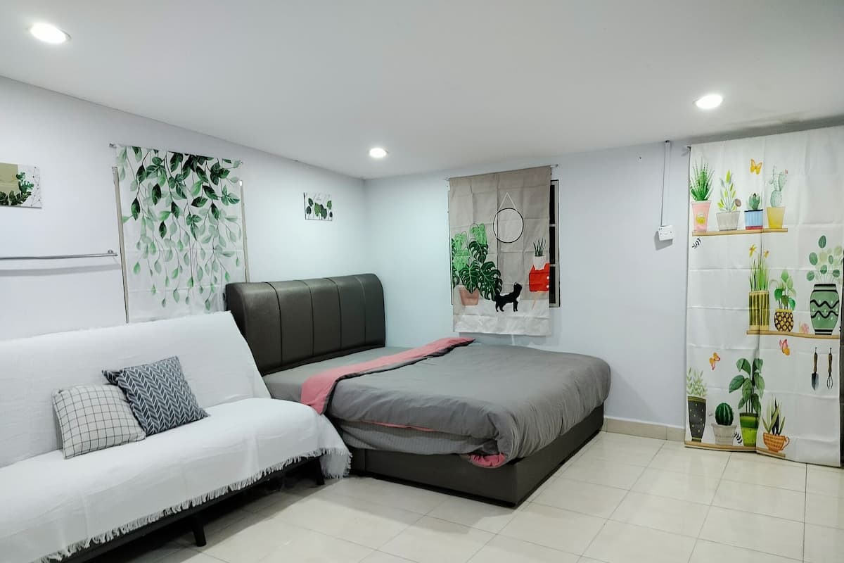 Une chambre moderne avec un canapé gris, un lit recouvert d’une couvre-lit grise et rose, et un mur décoré de rideaux à motifs végétaux.