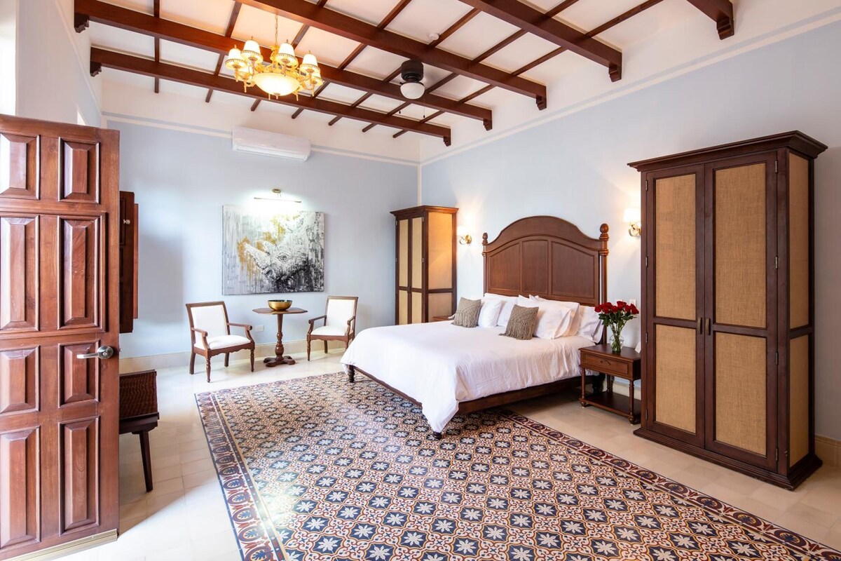 A bedroom with a large bed, a wooden wardrobe, a chandelier, a painting, and a patterned rug.