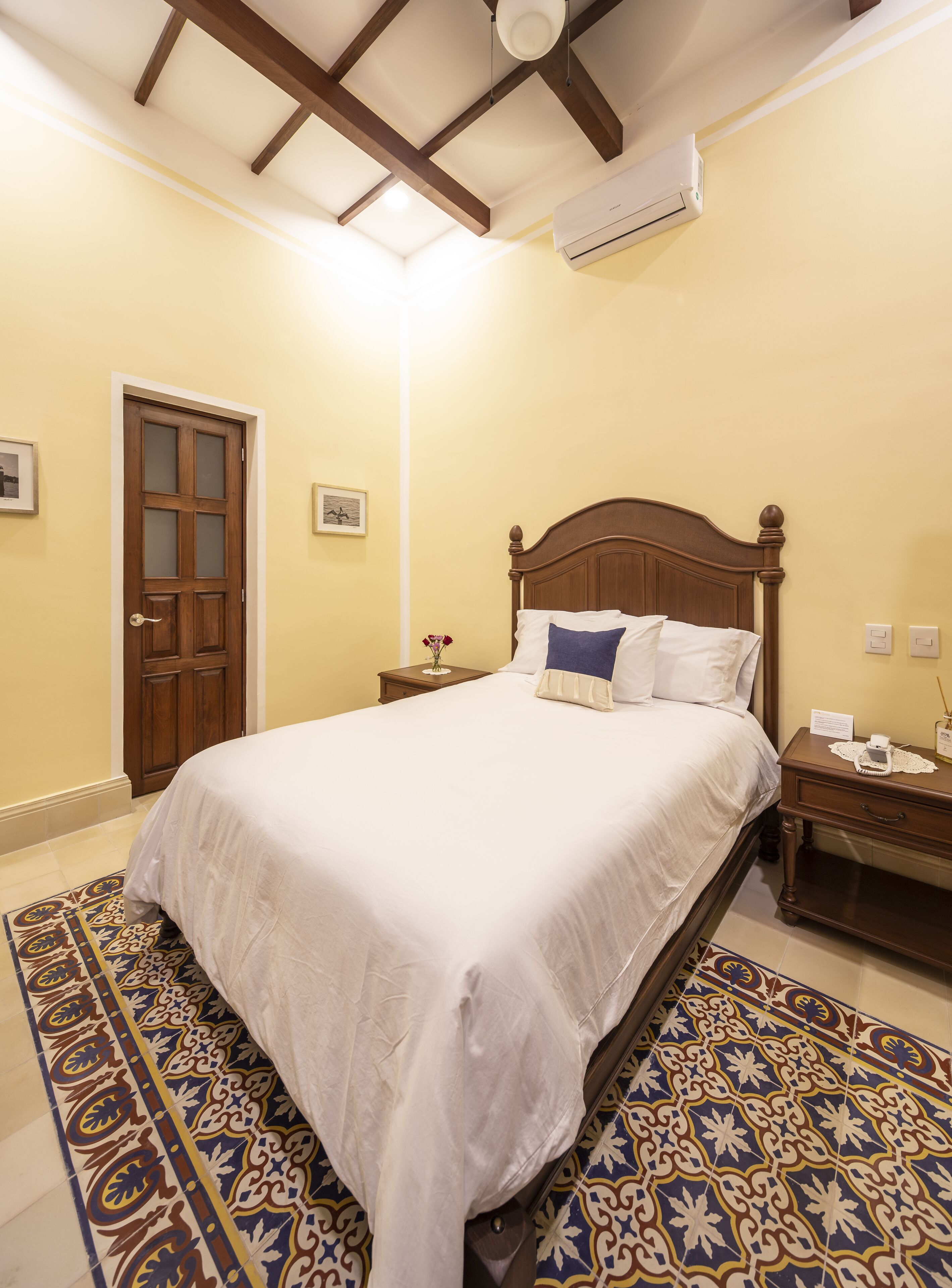 A neatly made bed in a room with a wooden ceiling, a patterned rug, and a door with glass panels.