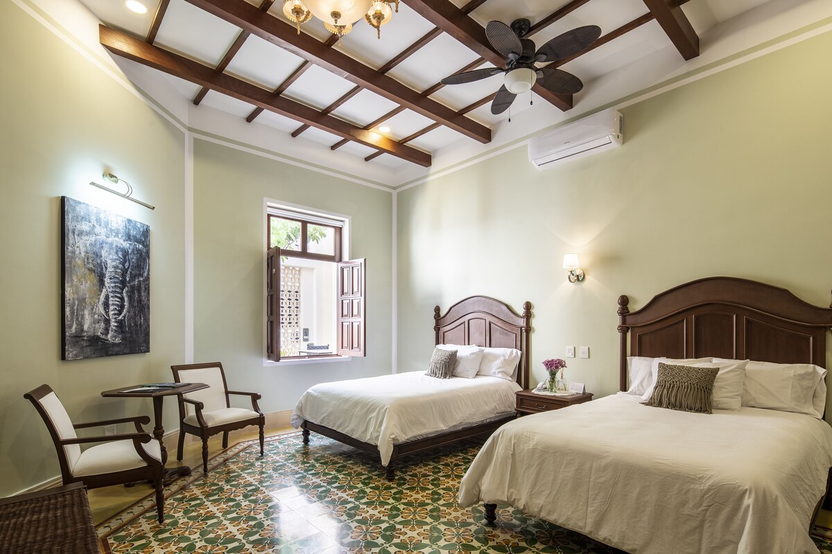A hotel room with two beds, a ceiling fan, a window with a view, a painting on the wall, and a tiled floor.