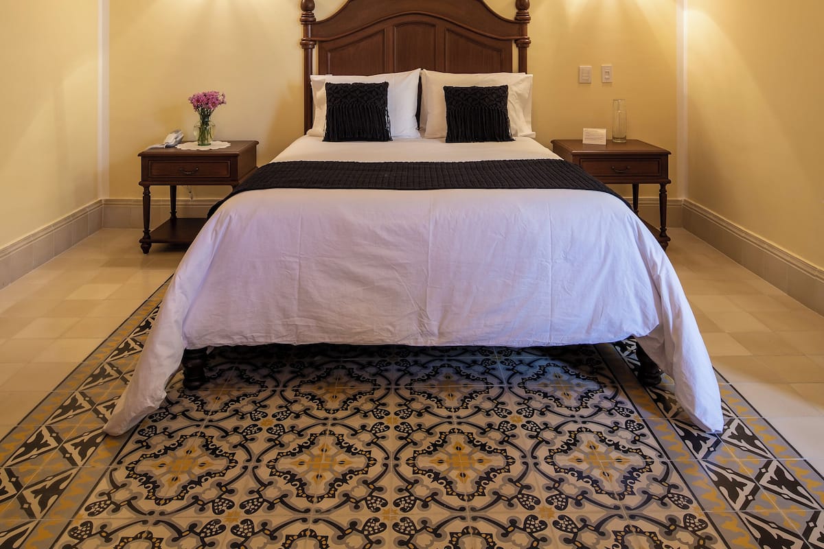 A hotel room with a large bed, wooden ceiling beams, two bedside lamps, and a patterned floor.