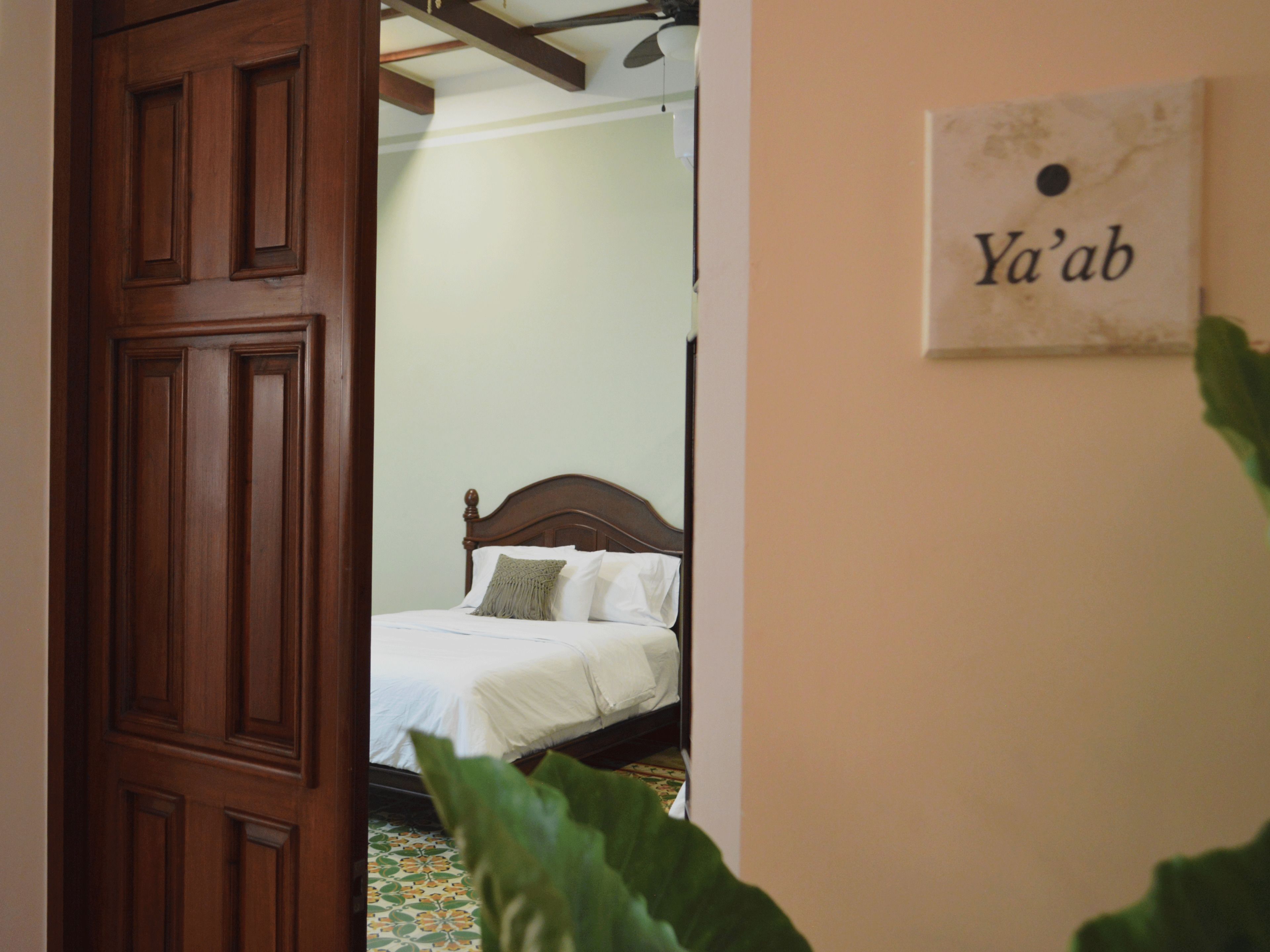 Un dormitorio con una cama de madera, una planta verde en primer plano y un letrero en la pared que dice "Ya'ab".