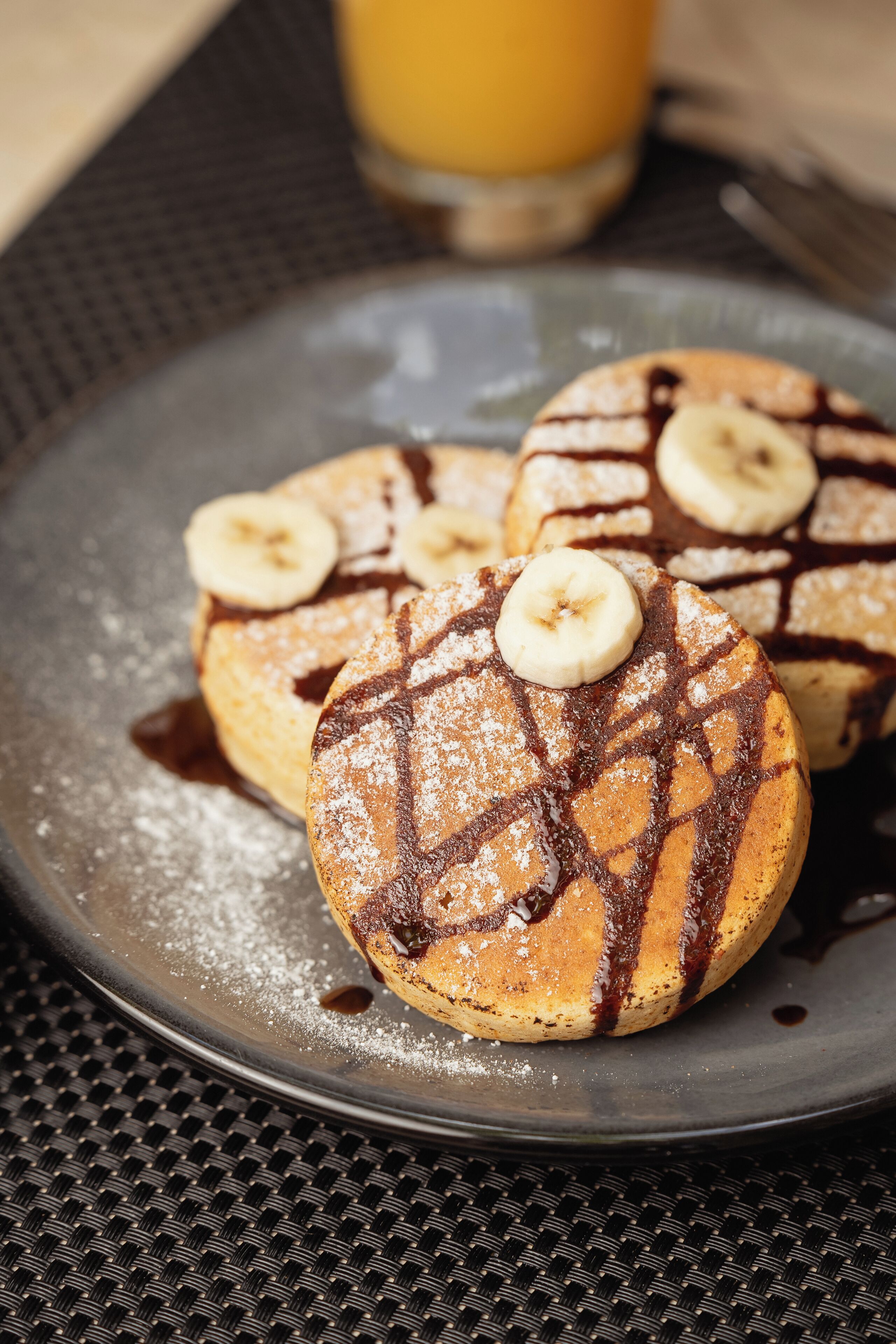A plate with pancakes topped with banana slices and chocolate syrup, dusted with powdered sugar.