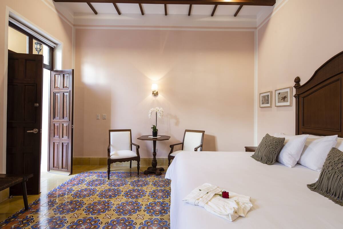 A hotel room with a bed, two chairs, a small table with a lamp, and a decorative wall.