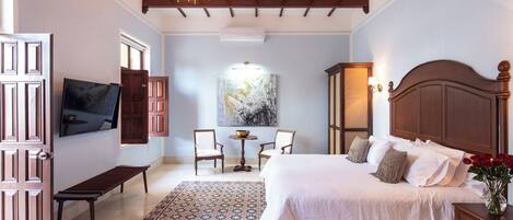 A bedroom with a large bed, a bench, two chairs, a table, and a TV on the wall.
