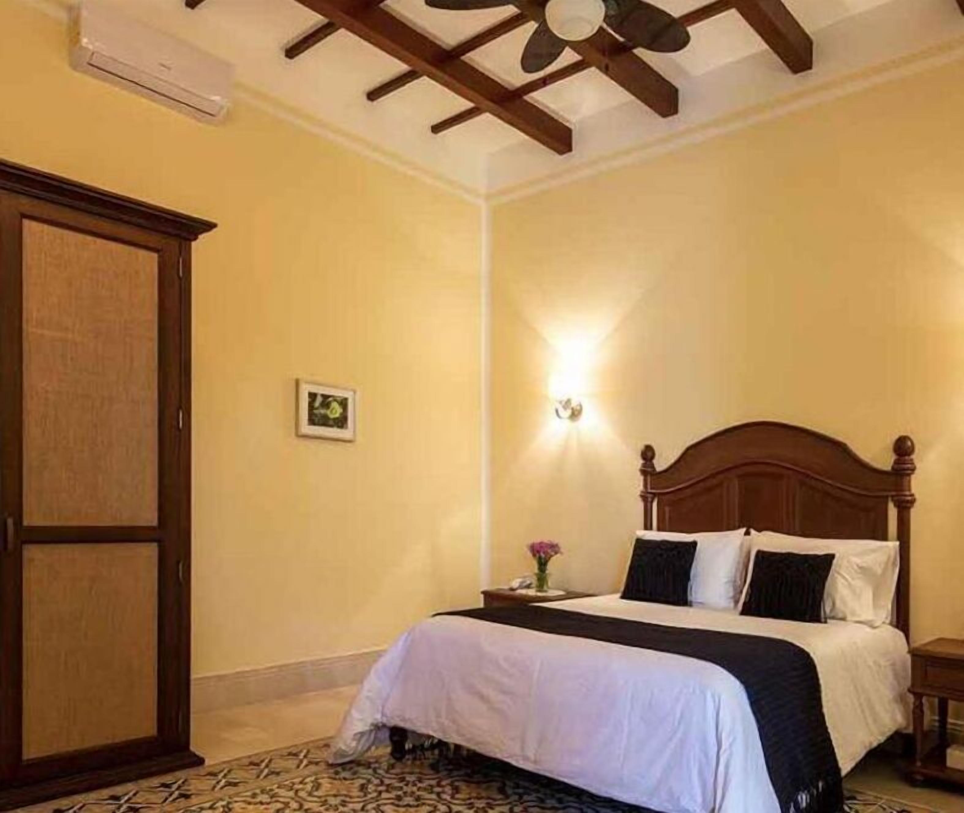 A hotel room with a wooden bed, a nightstand, a wall-mounted light, and a ceiling fan.