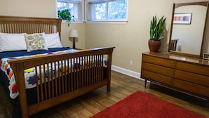 Ein Schlafzimmer mit einem hölzernen Bett, einem roten Teppich, einer Kommode, einem Spiegel und zwei mit Vorhängen versehenen Fenstern.