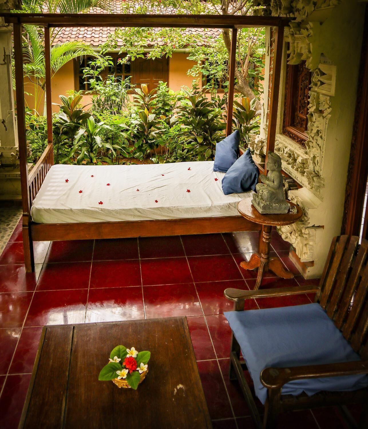 Una habitación con una cama de madera, una mesita con un arreglo floral, una silla azul y vistas a un jardín con diversas plantas.