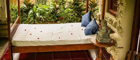 Una habitación con una cama de madera, una mesita con un arreglo floral, una silla azul y vistas a un jardín con diversas plantas.