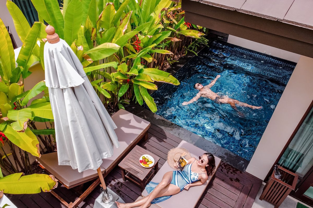 A pool area with a person swimming, a lounge chair, a table with a plate of fruit, and lush greenery.
