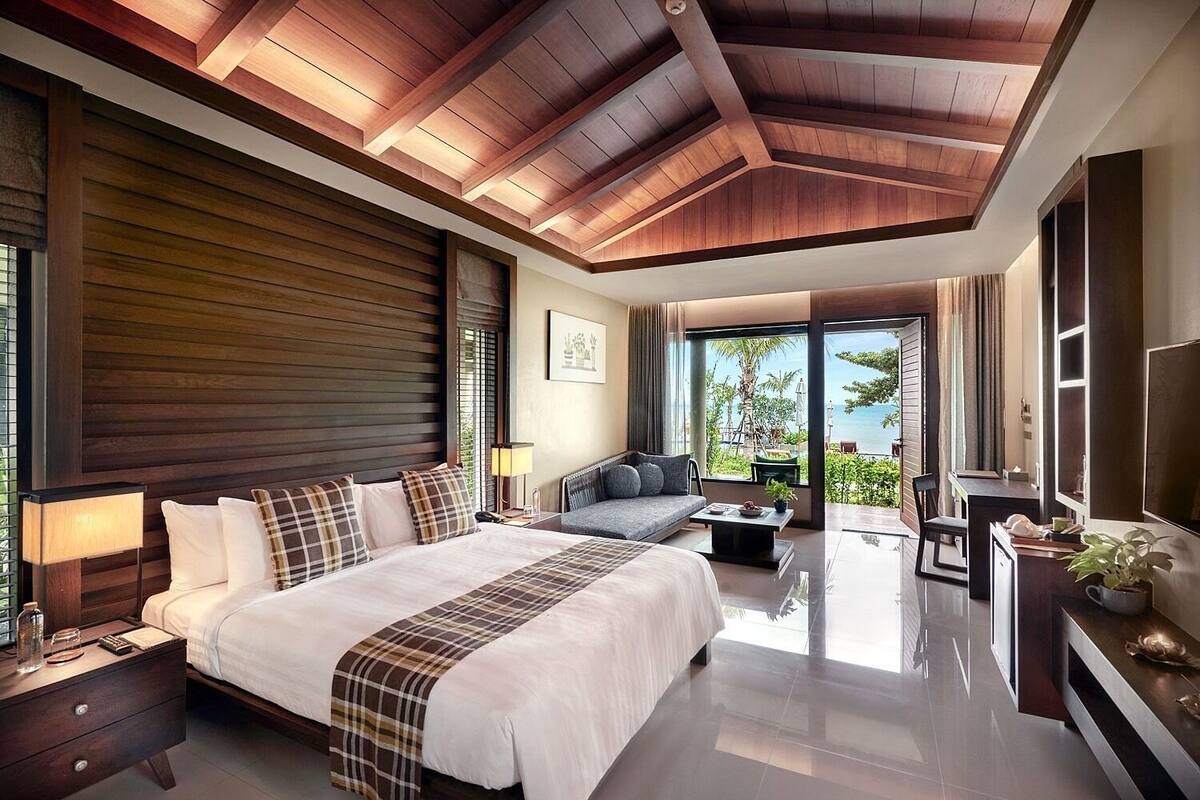 A modern hotel room with a large bed, a sofa, and a desk. The room has a wooden ceiling and large windows offering a view of the ocean.