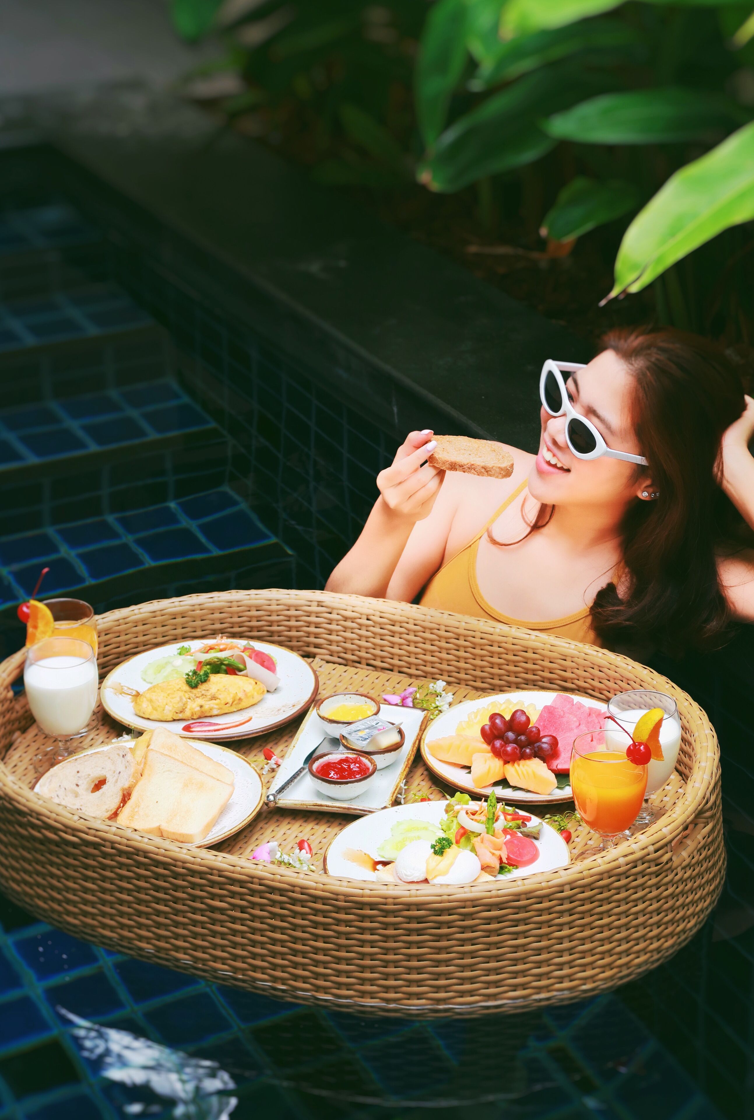 A wicker tray with a variety of breakfast items including toast, eggs, fruit, and a drink, placed on a tiled surface near a pool.