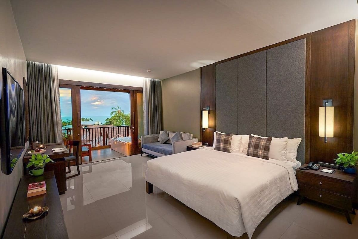 A hotel room with a large bed, a desk with a chair, a TV, and a balcony with a view of the ocean.