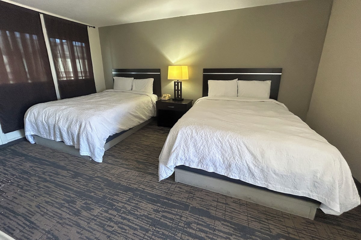 A hotel room with two single beds, a nightstand with a lamp, and a large window with curtains.
