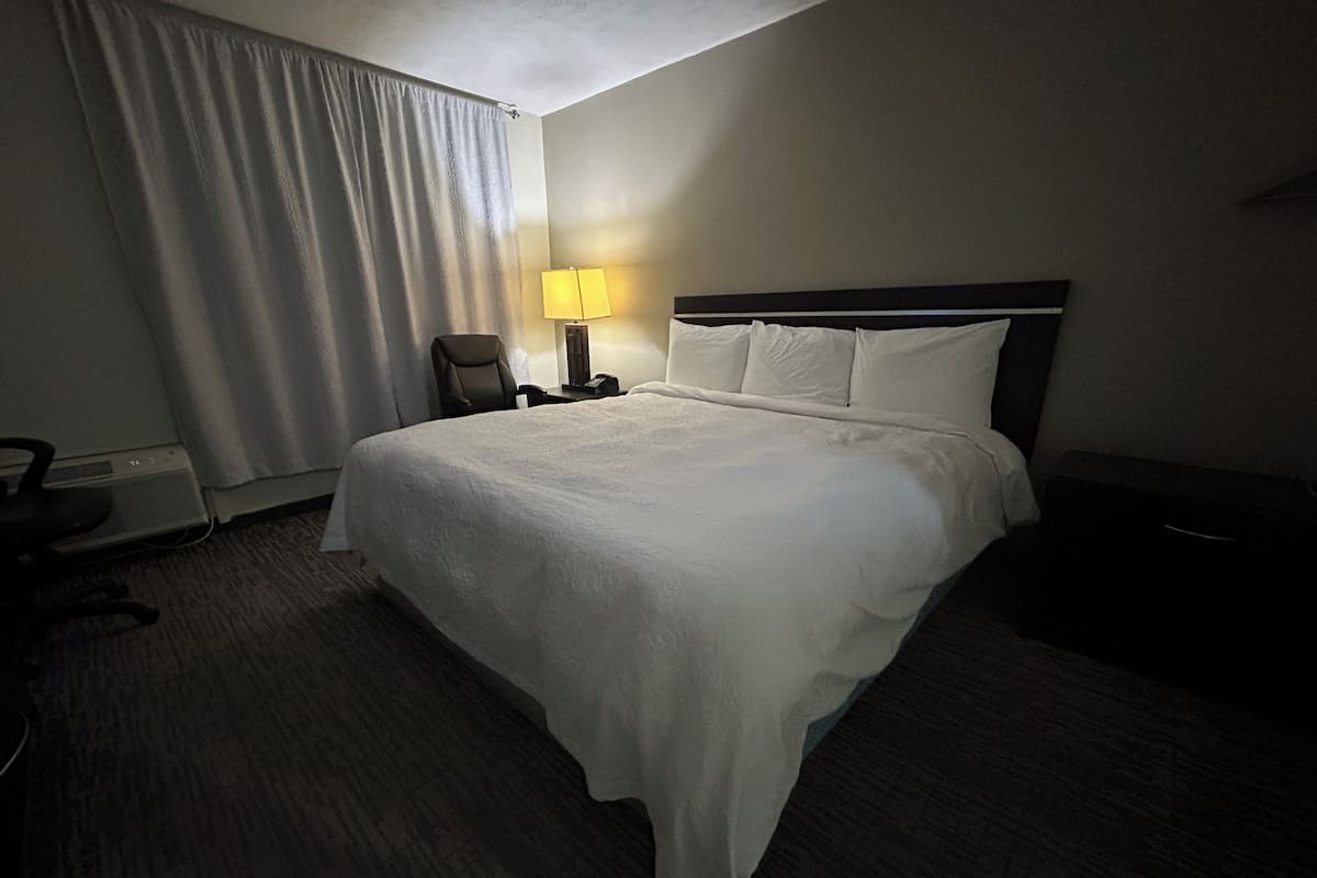 A hotel room with a large bed, a desk with a lamp, a chair, and a window with curtains.